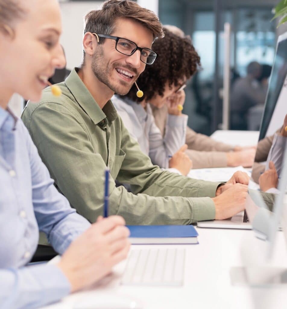 Professional customer support agent man in assisting customers at call center office, with colleagues in the background