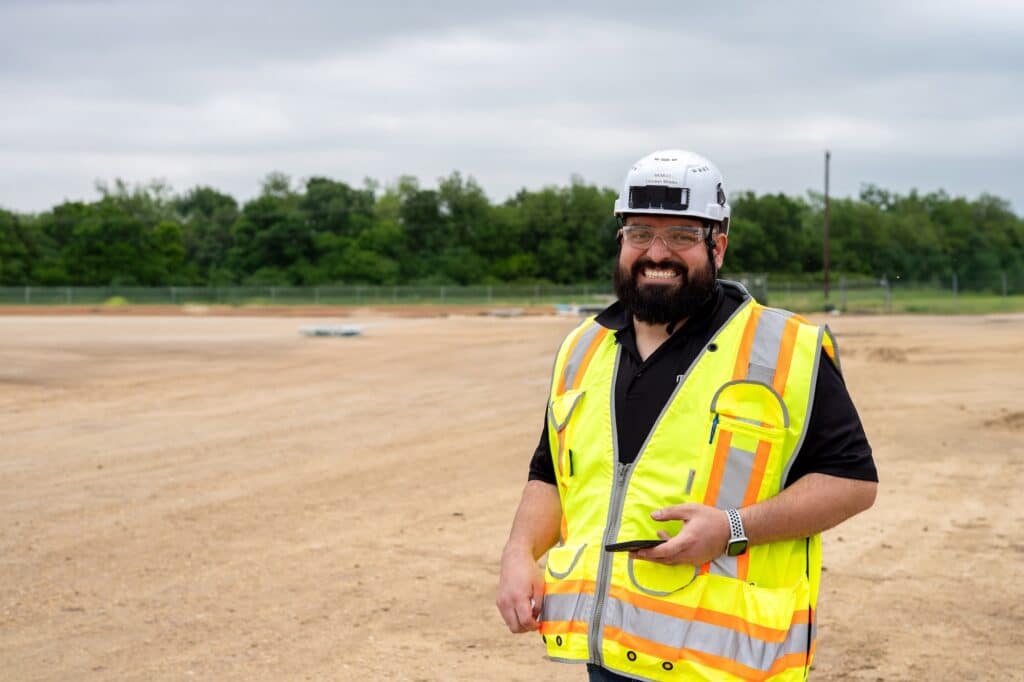A man wearing a hard hat and reflective safety vest stands smiling at a construction site with dirt ground and trees in the background.