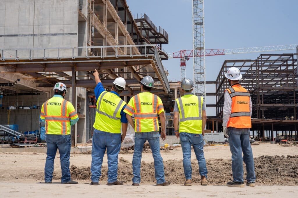 Five construction workers wearing safety vests and hard hats stand on a dirt site, looking at and discussing a building under construction with steel framework and a crane in the background.
